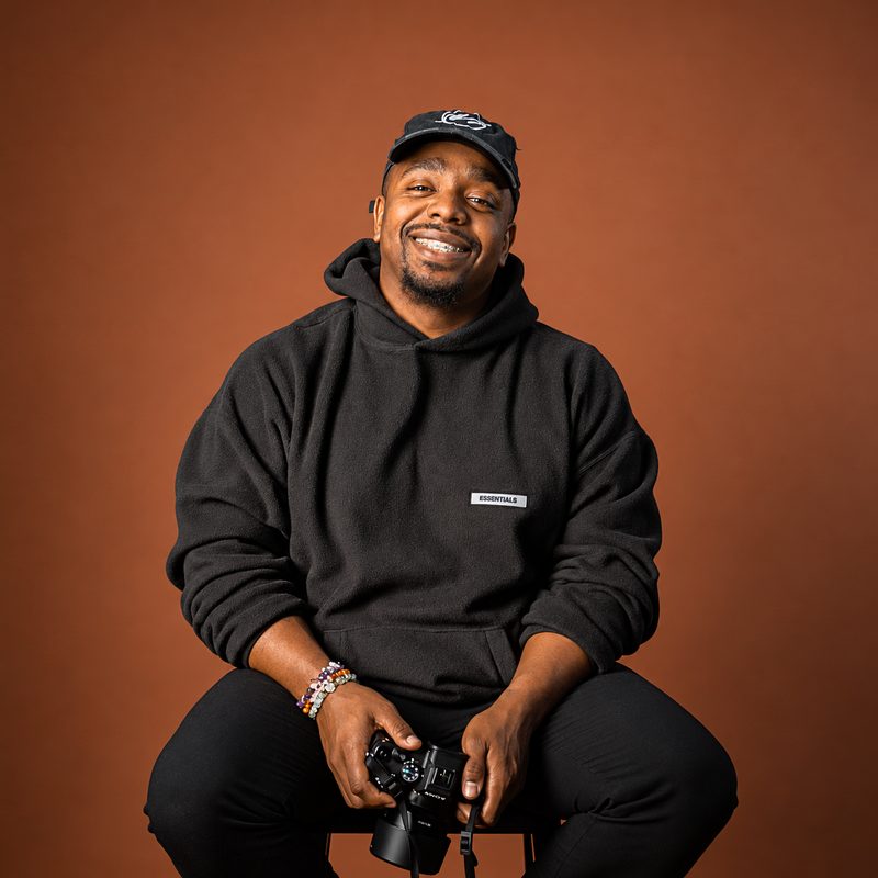 Tyquin Briggs smiling, seated on a stool in a black hoodie and cap, holding a Sony camera against a rust-orange backdrop