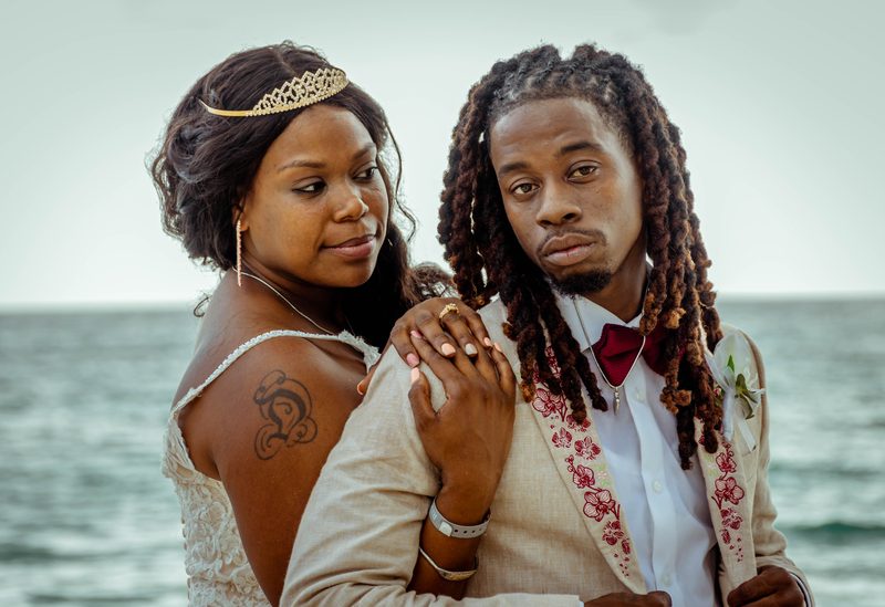Bride in a tiara resting her hand on the shoulder of a groom with locs and an orchid-embroidered jacket, the sea behind them