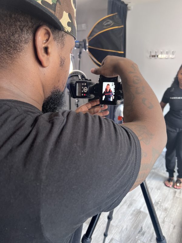 The photographer, seen over his shoulder, holding up a camera whose screen shows the woman standing in front of him in a studio with a softbox