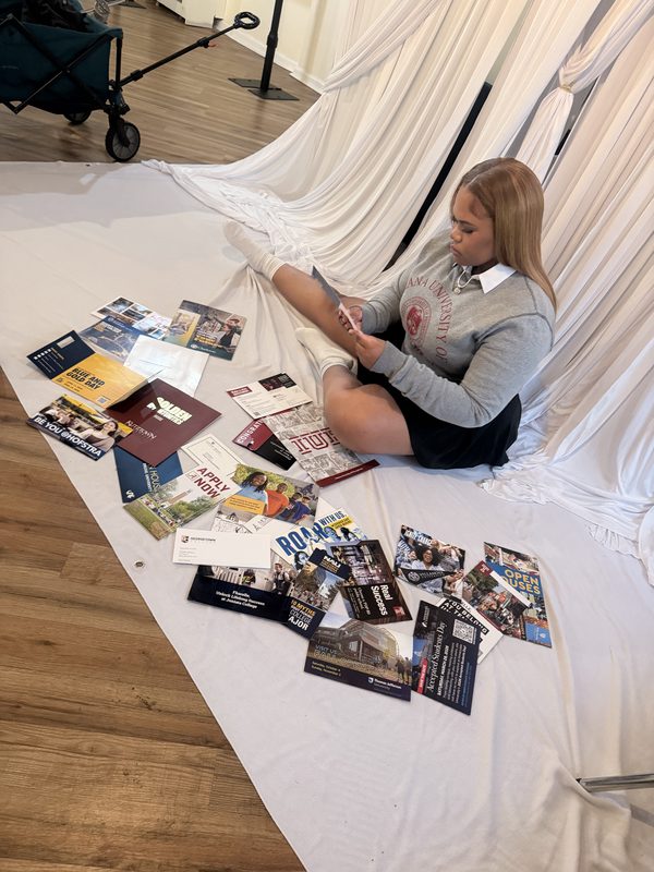 A graduate in a gray sweater sits on a white drape surrounded by college brochures and papers, arranged as a set