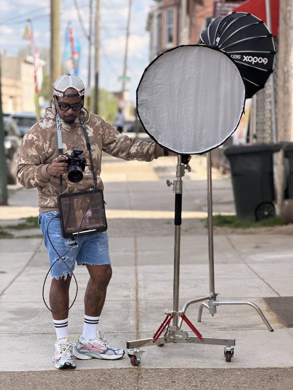 Tyquin Briggs on a city sidewalk holding a camera with a monitor, beside a large softbox and reflector on a stand
