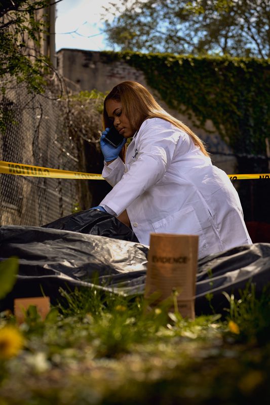 Model in a white medical examiner's coat and blue gloves on a phone, crouched over a black tarp beside an evidence bag in a staged alley set