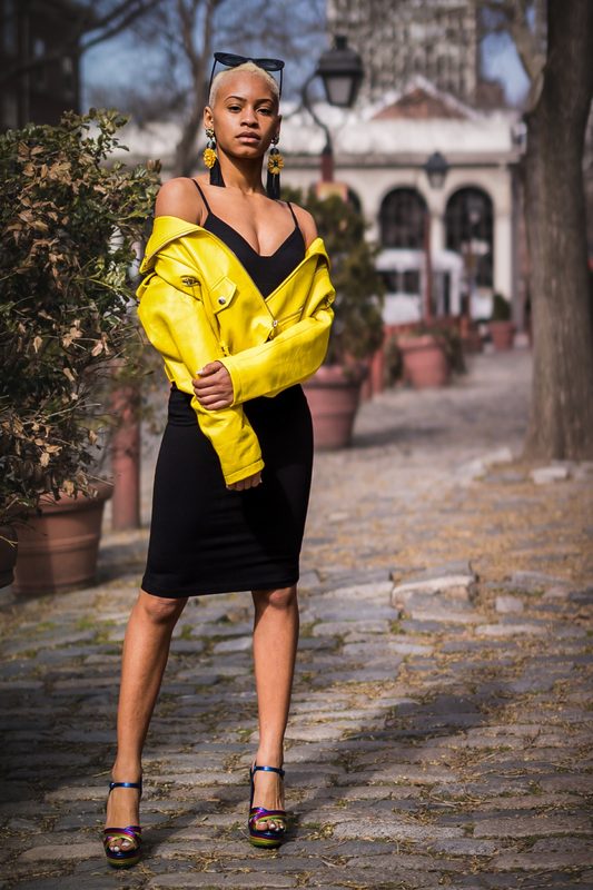 Woman in a yellow jacket and black dress standing on a brick sidewalk in front of historic buildings
