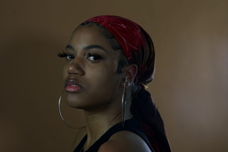 Woman in a red silk head wrap and large silver hoop earrings looking back over her shoulder against a warm brown backdrop