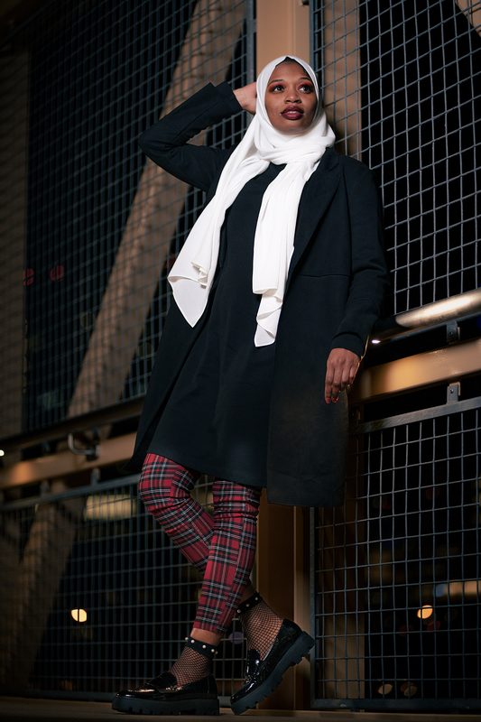 Woman in a black coat, white scarf, and plaid pants standing on a steel bridge